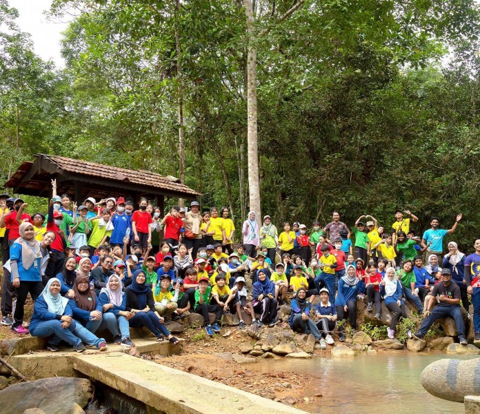 A large group of schoolchildren and researchers sit and stand together on the water edge with trees behind, waving and smiling at the camera